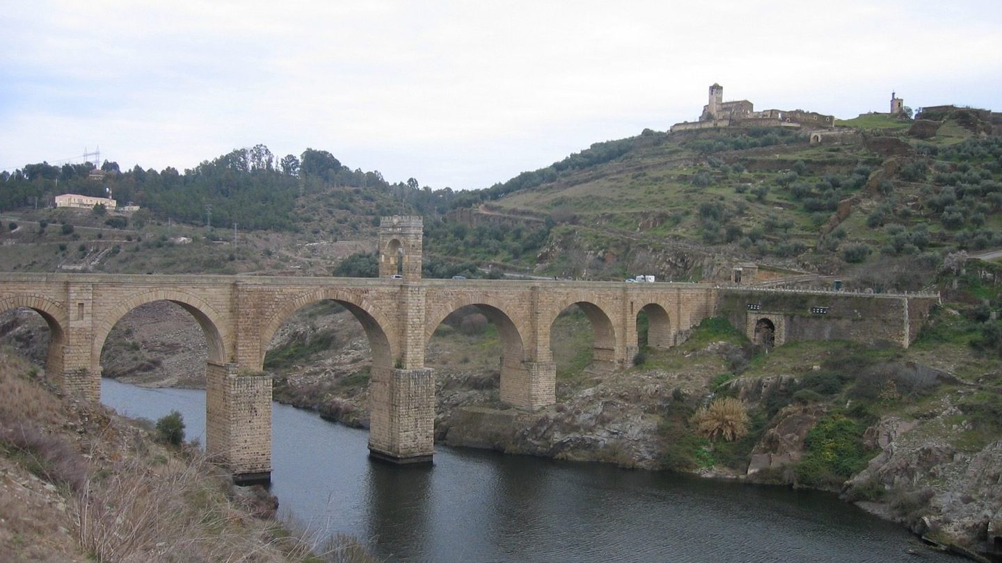 The Alcántara Bridge, a Roman stone arch bridge over the Tagus River in Spain, showing its full six-arch span with the central triumphal arch tower and a hilltop temple in the background