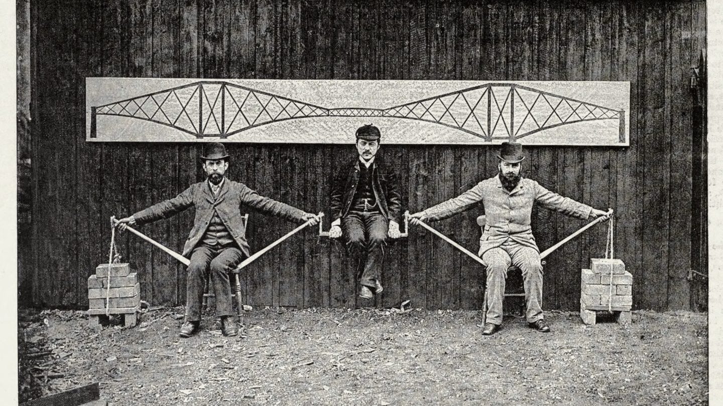 A circa-1890 postcard photograph of Benjamin Baker's human cantilever bridge model: two men seated on chairs with outstretched arms supported by poles and brick piles, with a third man suspended between them representing the load on the bridge's central span