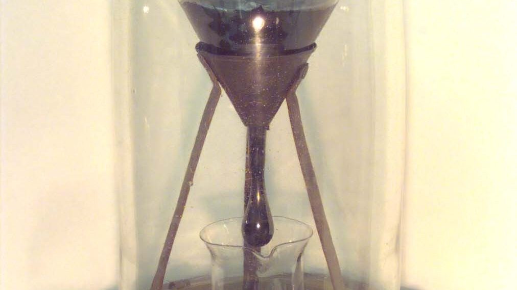 The University of Queensland's pitch drop experiment: a dark lump of pitch inside a sealed glass funnel, photographed with a 9-volt battery for scale