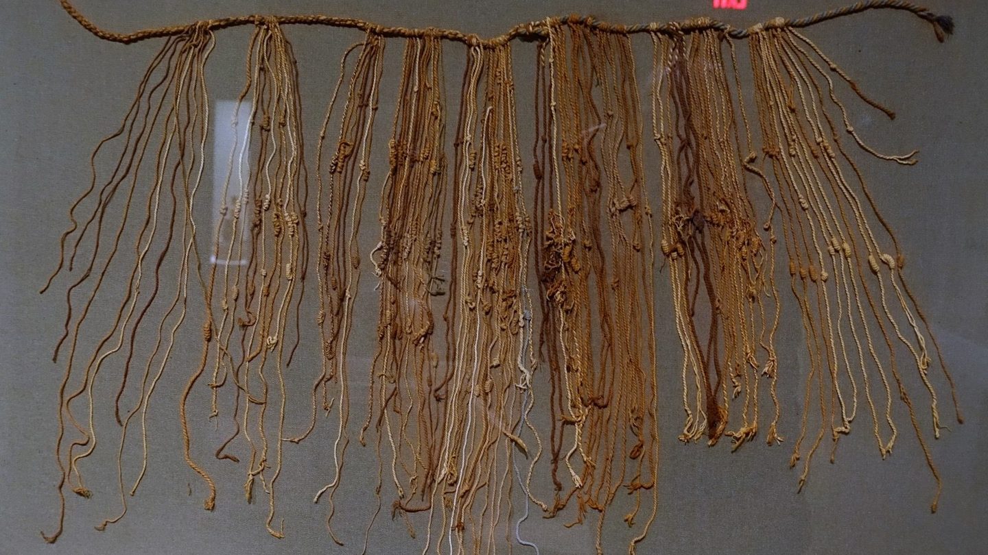 A fragmentary Inca quipu from the Late Horizon period, showing a thick horizontal main cord with dozens of thinner knotted pendant cords hanging from it in varied natural fiber colors